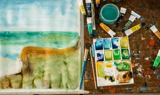 A colourful picture of painting, paint and brushes laid out on a table. On the left hand side is the painting and on the right hand side is different coloured paint a brushes, with a small pot of water. Underneath is a paint stained table.