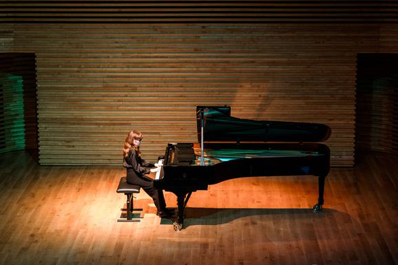 A young female sat at a piano in the middle of the stage. There is light shining down on the piano and her and there is a green light in some areas. She is looking forward a the piano and is dressed in black. The background is a dark brown wood colour.