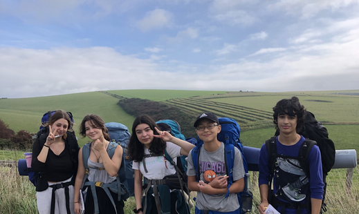 Group of young children standing on the top of a green hill, with large backpacks on. They are all smiling and facing the camera. There is blue sky and rolling hills in the background.